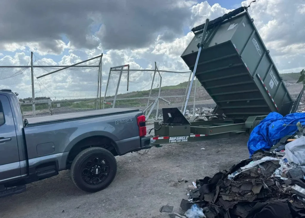 A gray pickup truck towing a small trailer with a raised green dumpster at a waste site, with a pile of debris and a blue tarp in the foreground and a chain-link fence in the background.