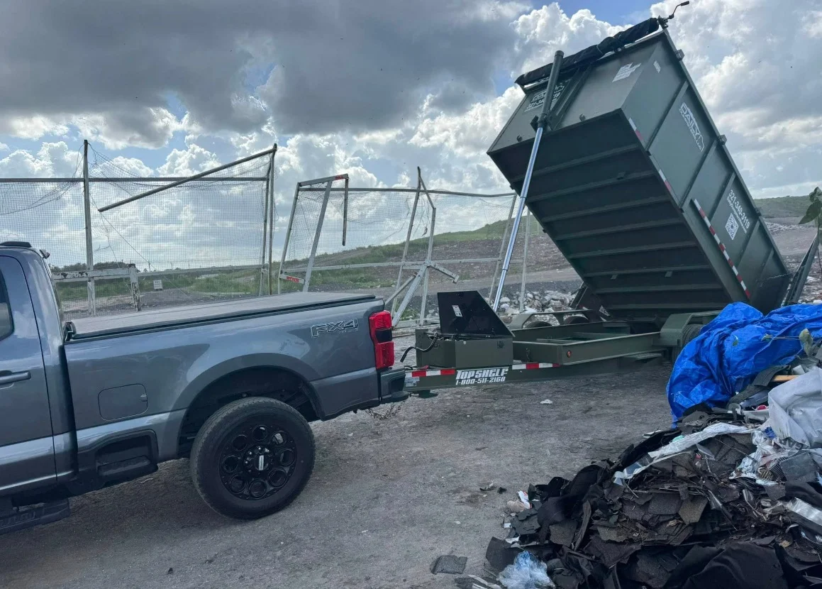 A gray pickup truck towing a small trailer with a raised green dumpster at a waste site, with a pile of debris and a blue tarp in the foreground and a chain-link fence in the background.