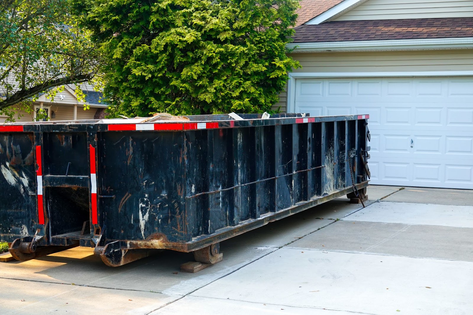A large black industrial dumpster sits on a residential concrete driveway in front of a white garage door.