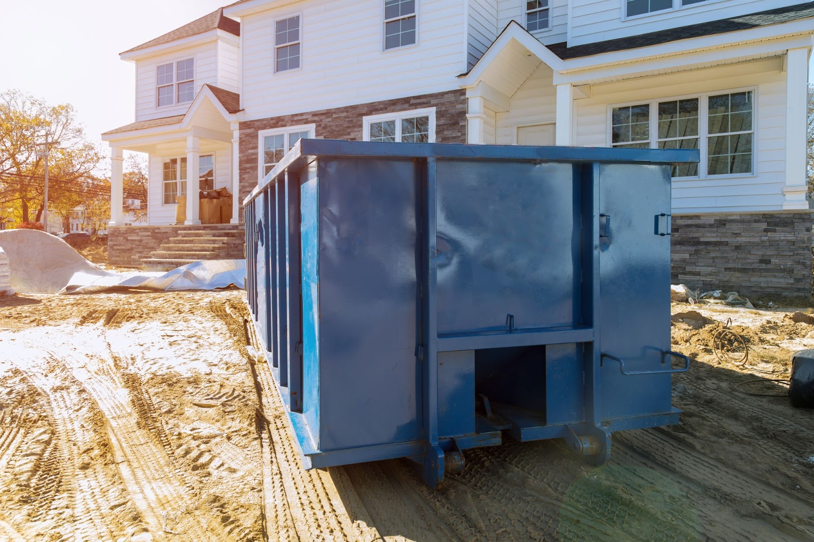 A large blue industrial dumpster sitting on a dirt construction site in front of a new white suburban house.