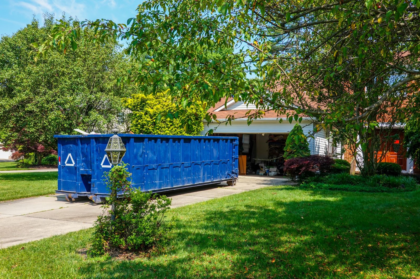 A large blue industrial dumpster sitting on a residential concrete driveway in front of an open white garage.